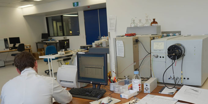 A person in a white lab coat working at a computer in a laboratory filled with scientific equipment.
