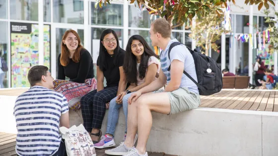 Group of five students chatting and smiling on a sunny campus plaza.