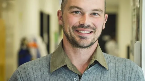 A smiling bald man wearing a collared shirt and standing in an office environment.