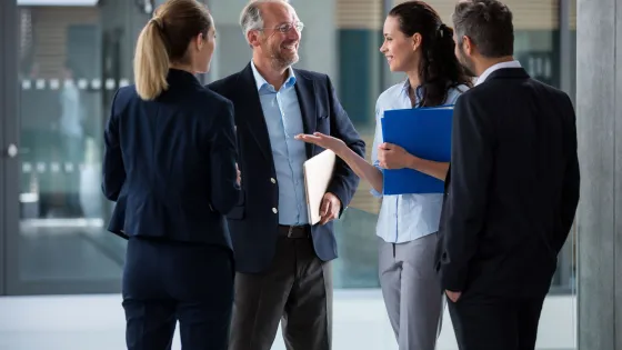 Four professionals are engaged in a conversation in an office hallway, with one man smiling and discussing something with three colleagues holding documents.