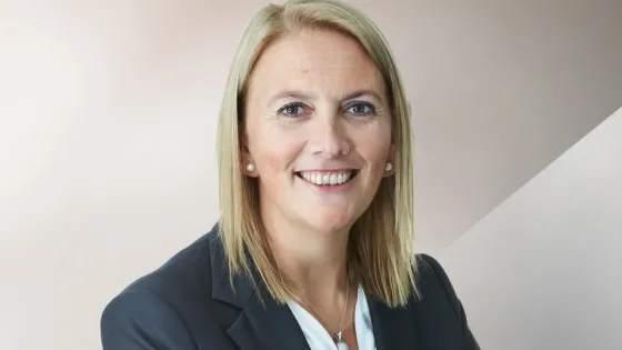 A professional headshot of a smiling woman with blonde hair, wearing a dark suit and white blouse, against a neutral, two-tone background.