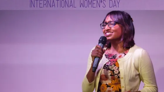 A woman with glasses speaks into a microphone at a "Science in the Pub" event on International Women's Day.