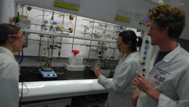 Three scientists wearing lab coats and safety glasses are working with chemical apparatus in a laboratory.